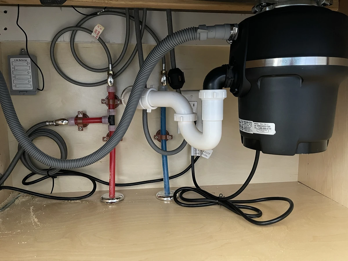 Under-sink garbage disposal install showing the full plumbing setup with red and blue PEX supply lines, shutoff valves, P-trap, dishwasher drain hose, and power cord by A.J. LeBlanc Heating in a Southern NH kitchen