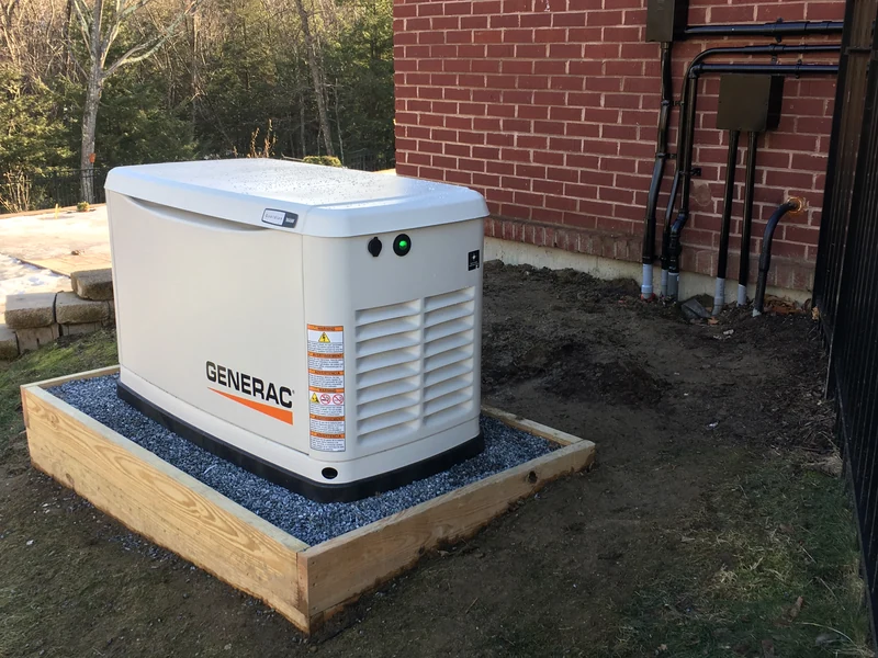 17kW Generac whole-home standby generator on a wood-framed gravel pad against a red brick Southern NH home, installed by A.J. LeBlanc Heating