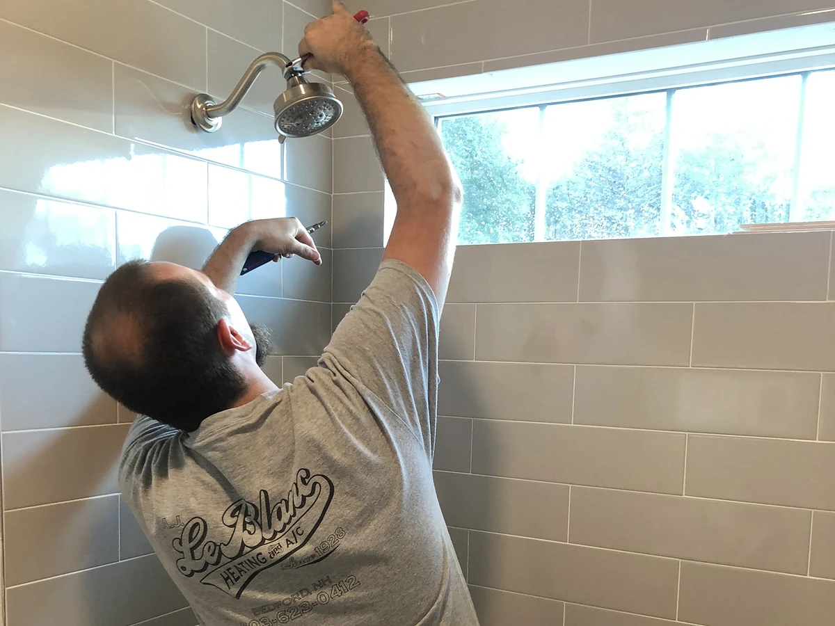 A.J. LeBlanc Heating licensed plumber installing a new brushed-nickel showerhead in a gray subway-tile shower at a Southern NH home