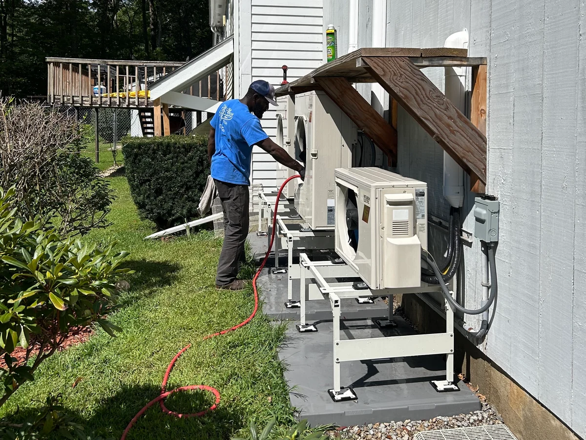 Technician cleaning outdoor mini split condenser units beside a home